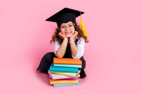 Full length photo of intelligent cheerful schoolkid wear mortarboard hat lean pile stack book empty space isolated on pink color background.