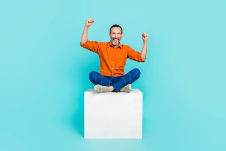 Photo of ecstatic man with white gray beard wear stylish shirt sit on cube raising fists up win bet isolated on teal color background.