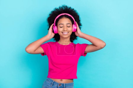 Photo of adorable relaxed girl with afro chevelure wear pink t-shirt arms touch headphones eyes closed isolated on blue color background.