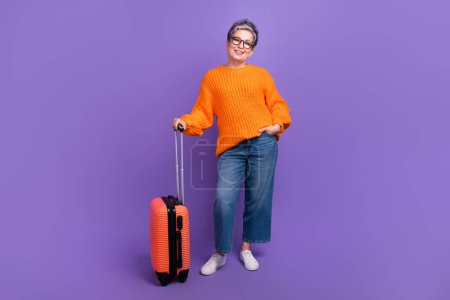Ganzkörper-Foto der jungen Model-Frau trägt orangefarbenen Strickpullover Jeans posiert mit Koffer isoliert auf lila Hintergrund.