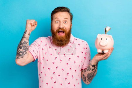 Portrait of delighted cheerful person raise fist hand hold money pig bank isolated on blue color background.
