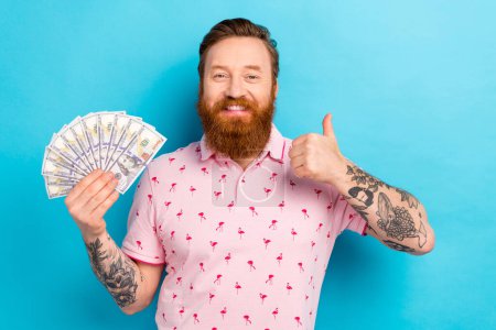 Photo de bel homme avec barbe rousse porter élégant t-shirt rose tenir dollars montrent pouce jusqu'à approbation isolé sur fond de couleur bleue.
