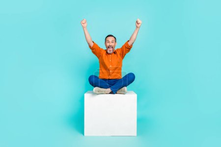 Full length photo of lucky excited man dressed orange shirt sitting platform rising fists isolated turquoise color background.
