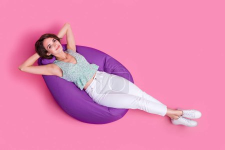 Full size photo of young charming smiling lady hands head take nap dreaming sleepy soft comfortable pouf isolated on pink color background.