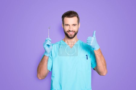 Portrait of joyful cheerful dental assistant in lab blue uniform gesturing thumbup approve advice yes done sign with finger, having tool in hand, isolated on grey background.