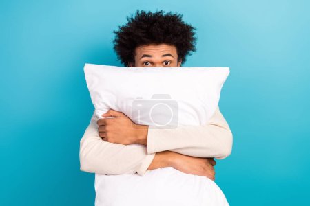 Photo portrait of nice young guy hugging pillow hide face wear trendy white pajama outfit isolated on blue color background.