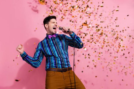 Portrait of carefree handsome guy arm hold wired microphone shout sing flying confetti isolated on pink color background.