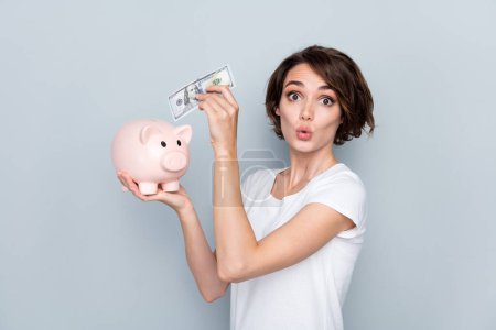 Portrait of funny funky playful girl bob hairdo wear stylish t-shirt put dollar in piggy box plump lips isolated on gray color background.