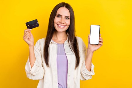 Photo of intelligent nice woman dressed white jacket demonstrate smartphone screen hold debit card isolated on yellow color background.