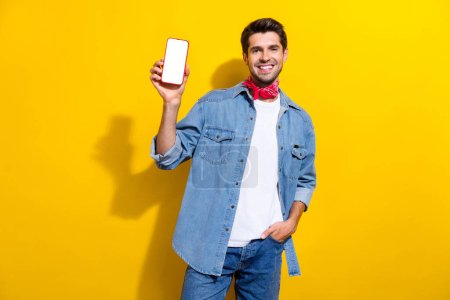Photo portrait of handsome young guy hold white screen device wear trendy jeans outfit isolated on yellow color background.