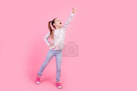 Ganzes Foto von guter Laune Mädchen tragen stilvolle Sweatshirt Jeans Arme sehen leer Raum erhobene Faust nach oben isoliert auf rosa Hintergrund.