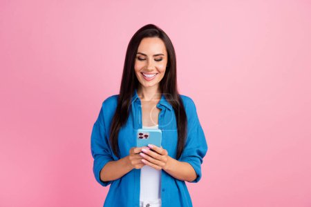 Porträt einer intelligenten Frau mit langen Frisuren trägt stylisches Hemd Blick auf Smartphone in den Händen im Chat isoliert auf rosa Hintergrund.