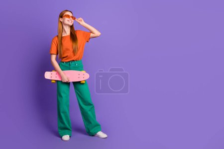 Photo de gaie fille mignonne porter des vêtements à la mode tenir skate look promo bannière nouvelles saisonnières espace vide isolé sur fond de couleur pourpre.