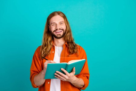 Portrait of intelligent writer with long hairstyle dressed orange shirt write novel with pen in hand isolated on teal color background.