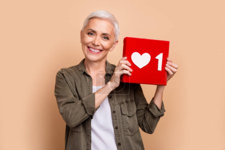 Foto de mujer de mediana edad sosteniendo botón rojo haga clic como icono en las redes sociales compartir contenido en youtube sobre fondo de color beige.