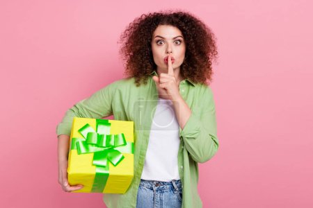Photo portrait of attractive young woman hold gift finger silence gesture dressed stylish green clothes isolated on pink color background.