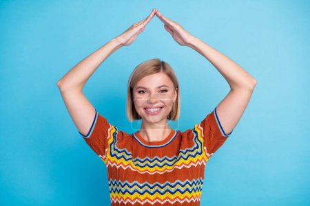 Portrait of positive cheerful girl with short hairstyle wear strited t-shirt arms making roof over head isolated on blue color background.