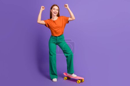 Photo de fille gaie positive porter des vêtements à la mode lever les poings oui gagner compétition équitation skate isolé sur fond de couleur pourpre.