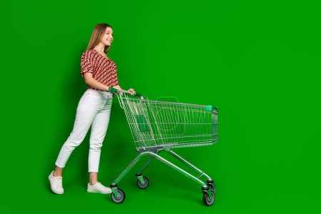 Foto de longitud completa de optimista adolescente vestido impresión blusa mirada espacio vacío en el supermercado con carro aislado sobre fondo de color verde.