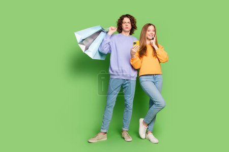Retrato de cuerpo completo de dos personas engañosa chica mostrar teléfono inteligente confundido chico mantenga bolsas de centro comercial espacio vacío aislado sobre fondo de color verde.