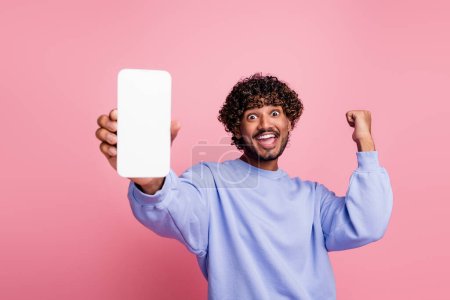 Photo portrait of handsome young male hold white screen raise fist winning wear trendy blue garment isolated on pink color background.