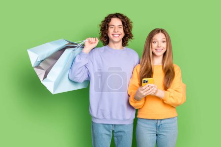 Retrato de dos personas adolescentes sonrisa dentada mantenga bolsas de centro comercial de la tienda de teléfonos inteligentes aislados en fondo de color verde.