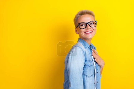 Photo portrait of lovely young lady specs look empty space touch chest dressed stylish denim garment isolated on yellow color background.