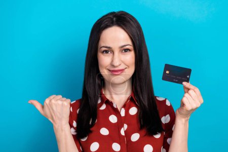 Photo portrait of lovely lady hold credit card point empty space dressed stylish dotted red garment isolated on blue color background.