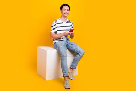 Full length photo of positive funky man wear striped t-shirt communicating modern device empty space isolated yellow color background.