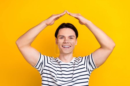 Photo of funny positive guy with brunet hair dressed striped t-shirt palms make roof over head isolated on yellow color background.