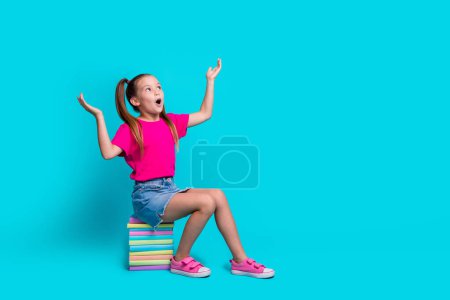 Full length photo of impressed schoolgirl wear t-shirt sit on book hold products staring at empty space isolated on teal color background.