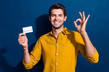 Photo portrait of attractive young man hold credit card show okey dressed stylish yellow clothes isolated on dark blue color background.