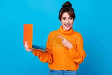Photo of excited pretty woman dressed orange sweater pointing finger modern device empty space isolated blue color background.