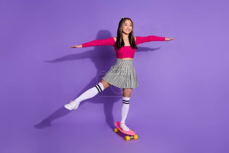 Full length photo of positive sweet girl wear pink top arms sides riding skateboard isolated purple color background.