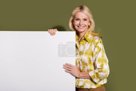 Porträt von fröhlich froh blonde Haare Promoter reifen Alters Frau hält Attrappe Informationstafel isoliert auf khakifarbenem Hintergrund.