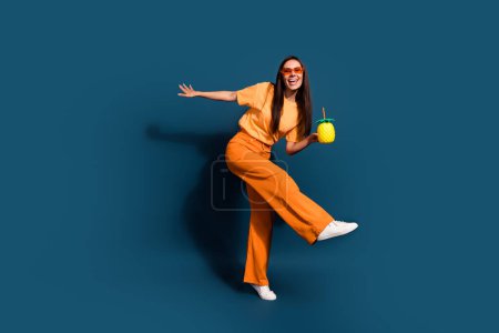 Tamaño de cuerpo completo foto joven dama en pantalones de color naranja y camiseta le gusta taza de piña cóctel de tequila en el resort sobre fondo de color azul oscuro.