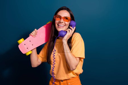 Portrait de femme hipster portant t-shirt orange appel vieux téléphone inviter des amis monter skateboard isolé sur fond de couleur bleu foncé.