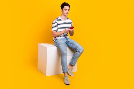 Full length photo of good mood cool guy dressed striped t-shirt chatting modern gadget empty space isolated yellow color background.