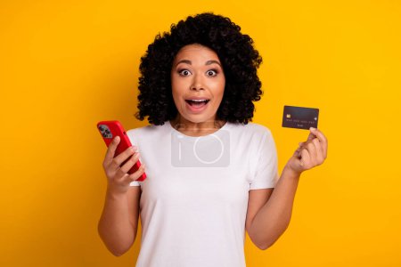 Photo de belle femme attrayante impressionnée avec chevelure tenant smartphone carte en plastique regardant isolé sur fond de couleur jaune.