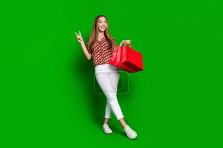 Foto de tamaño completo de adorable adolescente desgaste impresión blusa pantalones blancos mantenga bolsas muestran v-signo aislado en el fondo de color verde.