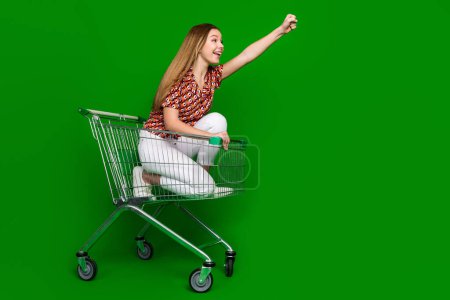 Foto de tamaño completo de la chica impresionada emocionada cabalgando en el carrito de la compra levantando el puño hacia arriba mirar espacio vacío aislado sobre fondo de color verde.