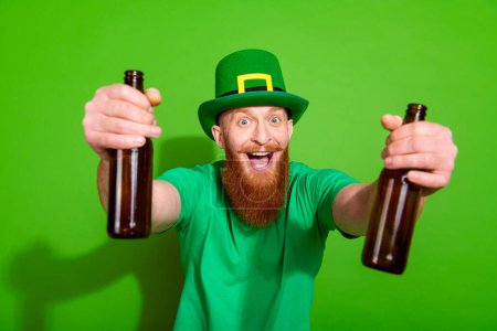 Portrait of cheerful funky young man open mouth hands hold glass beer bottles isolated on green color background.