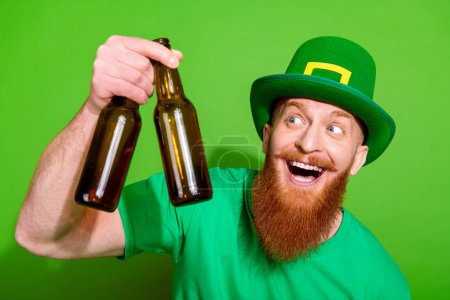Portrait of positive astonished person look arm hold glass beer bottles isolated on green color background.