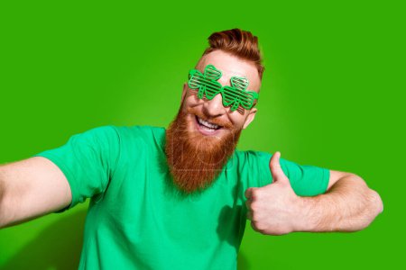Foto von attraktiven optimistischen Mann mit Ingwerbart in Kleeblättern zeigt Daumen nach oben Make-Selfie isoliert auf grünem Hintergrund.