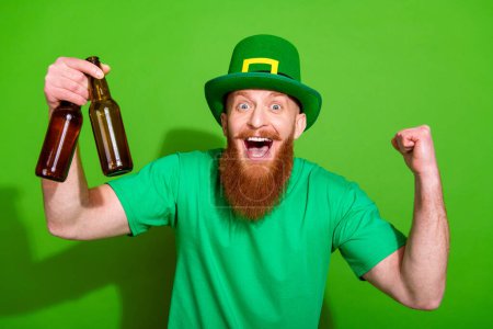 Photo of ecstatic delighted handsome man hold beer bottles raise fist isolated on green color background.