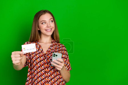 Photo de belle charmante fille habillée chemise d'impression tenant smartphone carte de crédit regarder espace vide isolé sur fond de couleur verte.