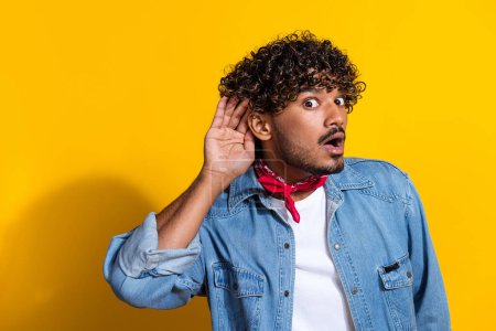 Photo portrait of attractive young man palm ear listen news dressed stylish denim clothes red scarf isolated on yellow color background.