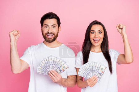 Foto-Porträt von attraktiven jungen Paar halten Banknoten Fan heben die Fäuste tragen trendige weiße Kleidung isoliert auf rosa Hintergrund.