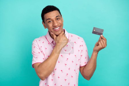 Portrait of positive minded guy hand touch chin hold plastic debit card isolated on turquoise color background.