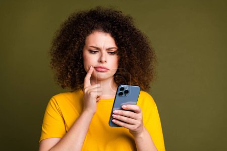 Portrait of young woman wearing yellow t shirt thoughtful browsing newsfeed touch cheek find trends isolated on khaki color background.